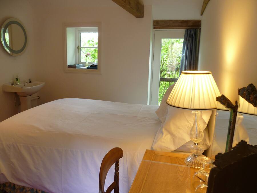 A bedroom with a bed, a bedside table with a lamp, a mirror, and a window with a view of greenery.