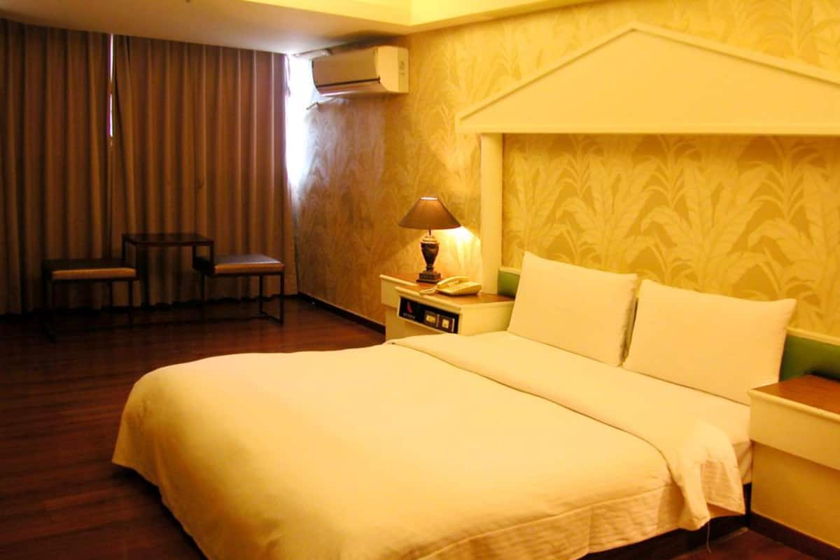 Habitación de hotel con una cama grande, una mesita de noche con lámpara, una silla y un equipo de aire acondicionado en la pared.