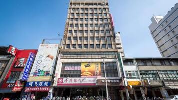 A multi-story building with a large billboard on the ground floor, several shops on the ground level, and a clear blue sky in the background.