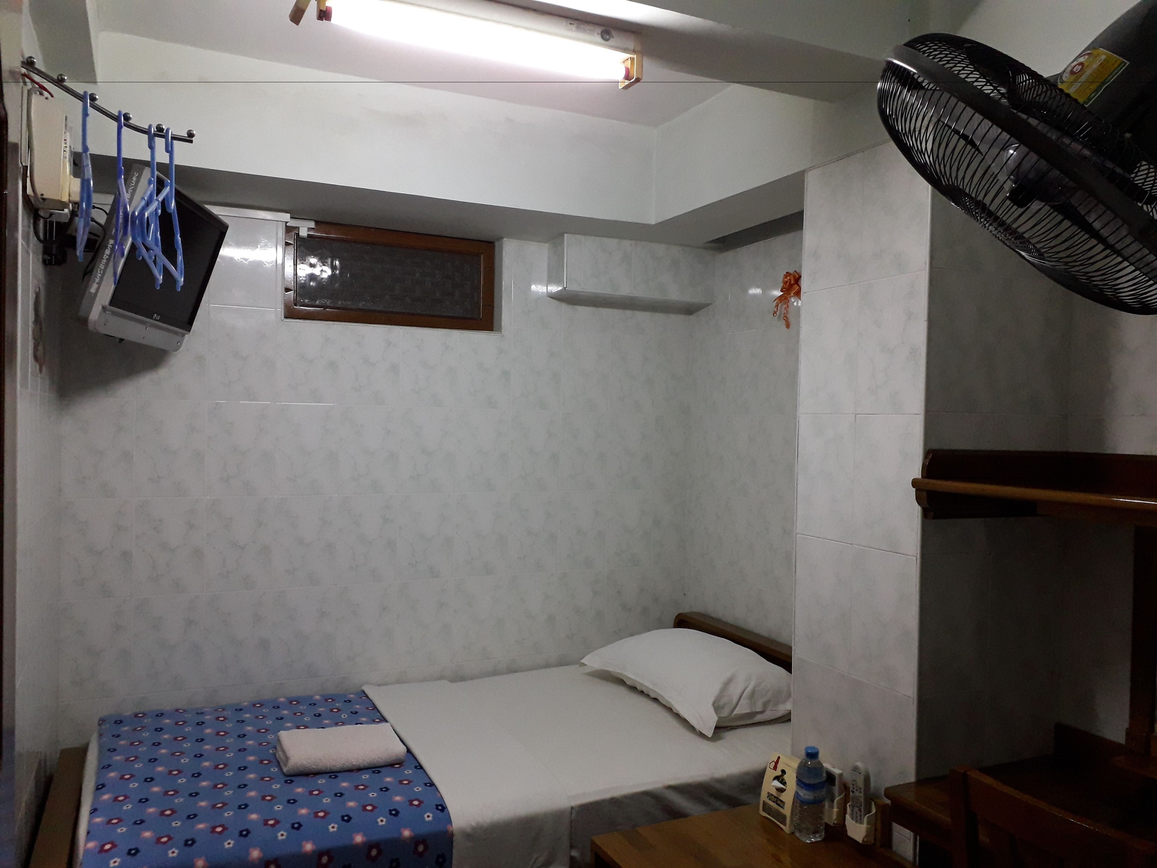 Una habitación pequeña y funcional con una cama individual, un escritorio, una silla, una lámpara de escritorio, un ventilador en la pared y un ventanal.