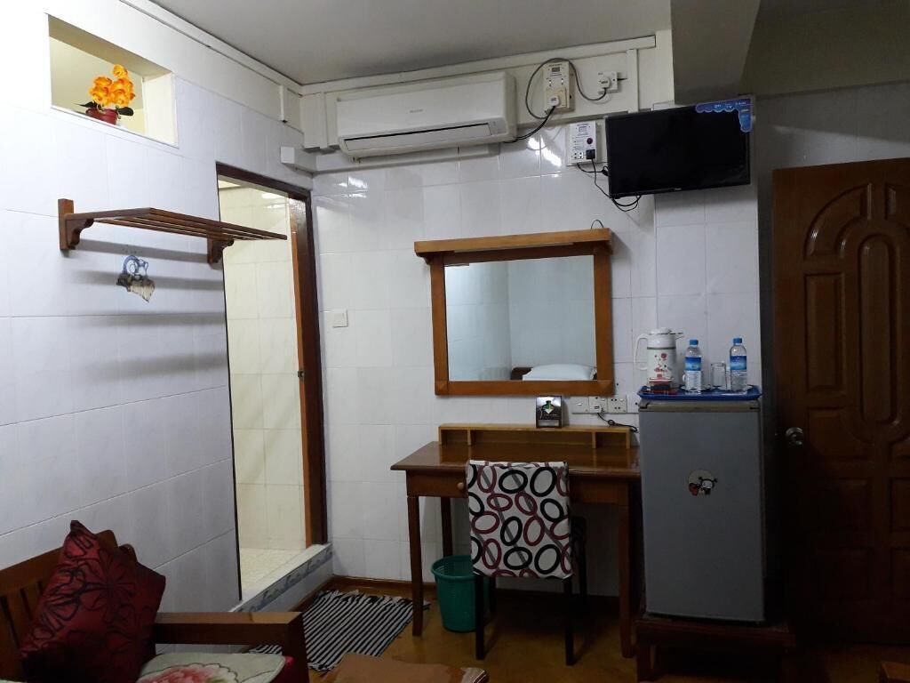 A compact hotel room with a wooden desk, a mirror, a small shelf with toiletries, a door, and a window with a flower vase.