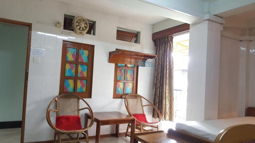 Una habitación con dos sillones de mimbre con cojines rojos, una mesa de madera, una cama, una ventana con cortinas y un aire acondicionado de pared.