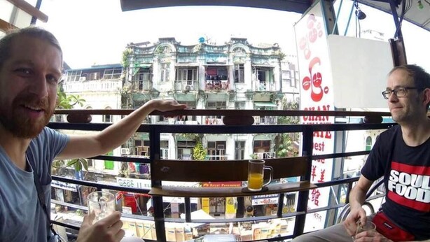 Two people sitting at a table with drinks, overlooking a street with buildings and shops. - Chan Myae Guest House (Yangon)