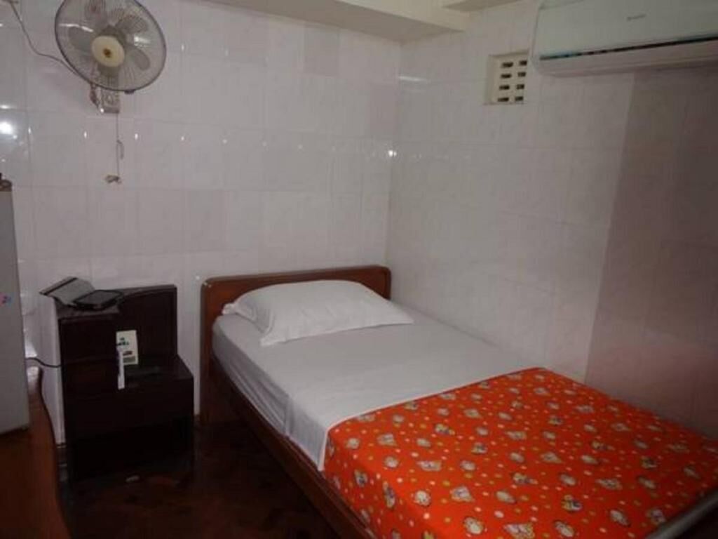 Une petite chambre avec un lit simple, dotée d’une tête de lit en bois, d’un oreiller blanc et d’une couverture rouge. On y trouve une table de chevet avec une lampe, ainsi qu’une petite table sur laquelle est posé un ordinateur portable. Un ventilateur de plafond est visible, et un appareil de climatisation est fixé au mur.
