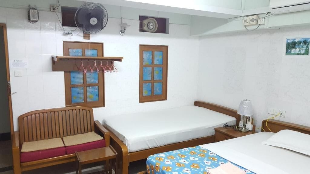 A room with a bed, a wooden bench, a small table, a bedside lamp, and two windows with informational posters.