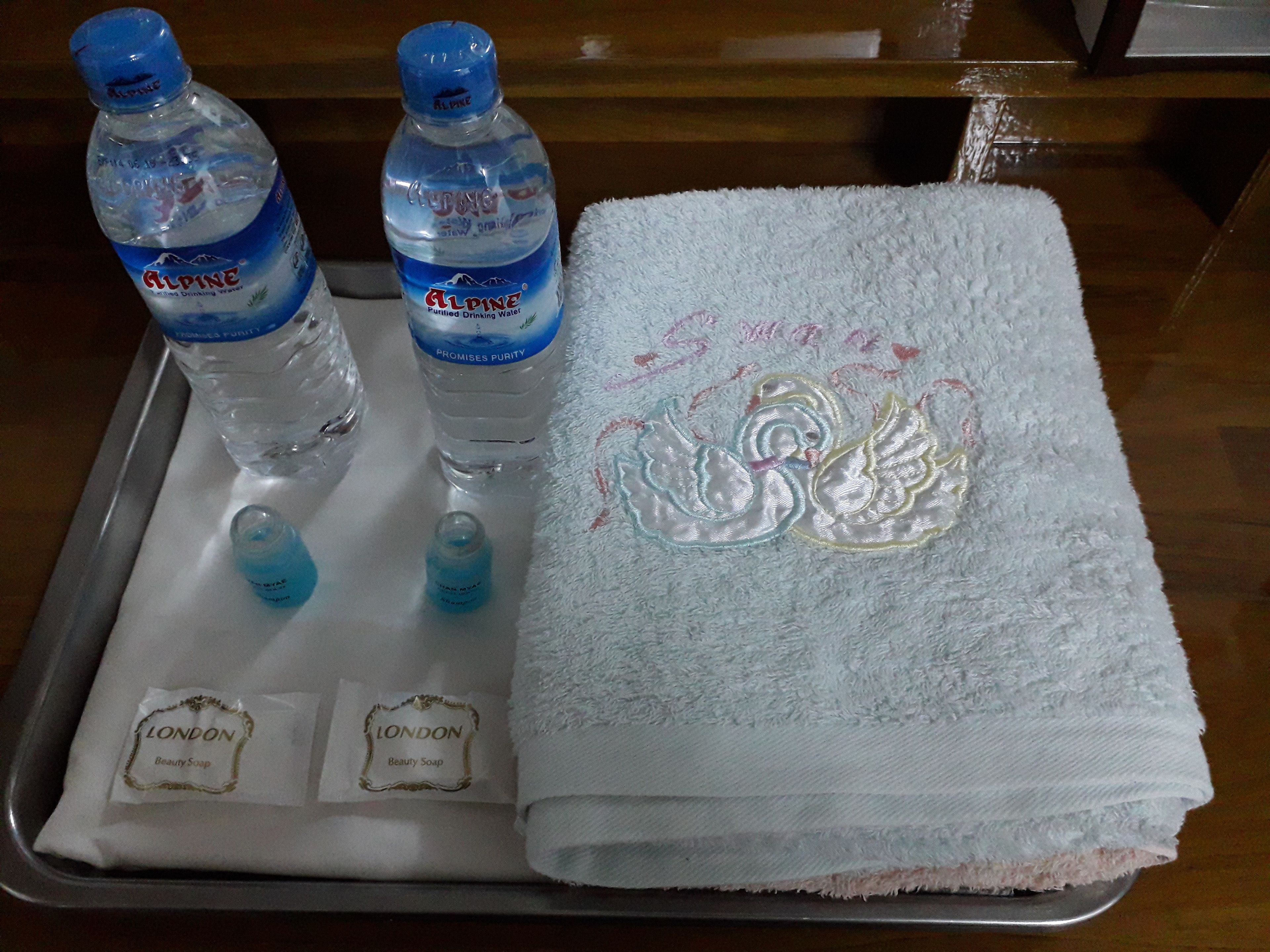 Una bandeja con dos botellas de agua Alpina, dos pequeñas botellas de líquido azul, una toalla blanca con bordado de cisne y dos pequeñas bolsitas con la etiqueta 'LONDON Beauty Soap'.