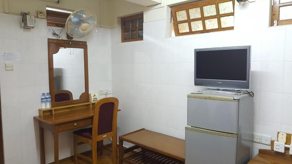 A compact room with a wooden desk, a chair, a small table, a refrigerator, a television, and a window with a view.