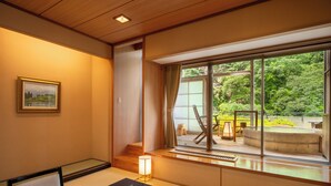 Ein traditioneller japanischer Raum mit Tatami-Boden, einem niedrigen Tisch, einem Stuhl, einer Lampe und einem großen Fenster mit Blick ins Grüne.