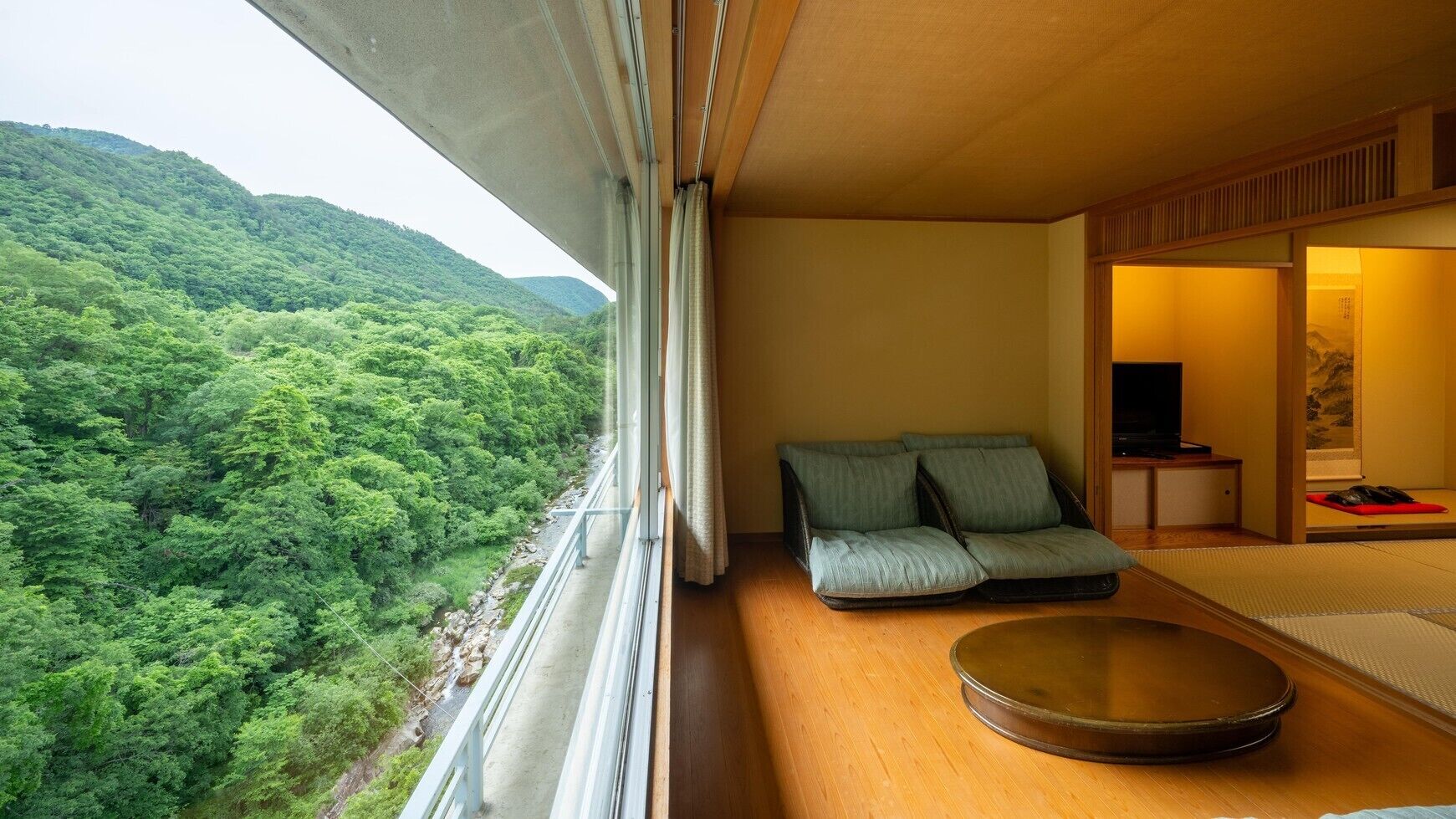 A room with a view of a forested hillside, a sofa, a tatami mat, and a small table with a television.
