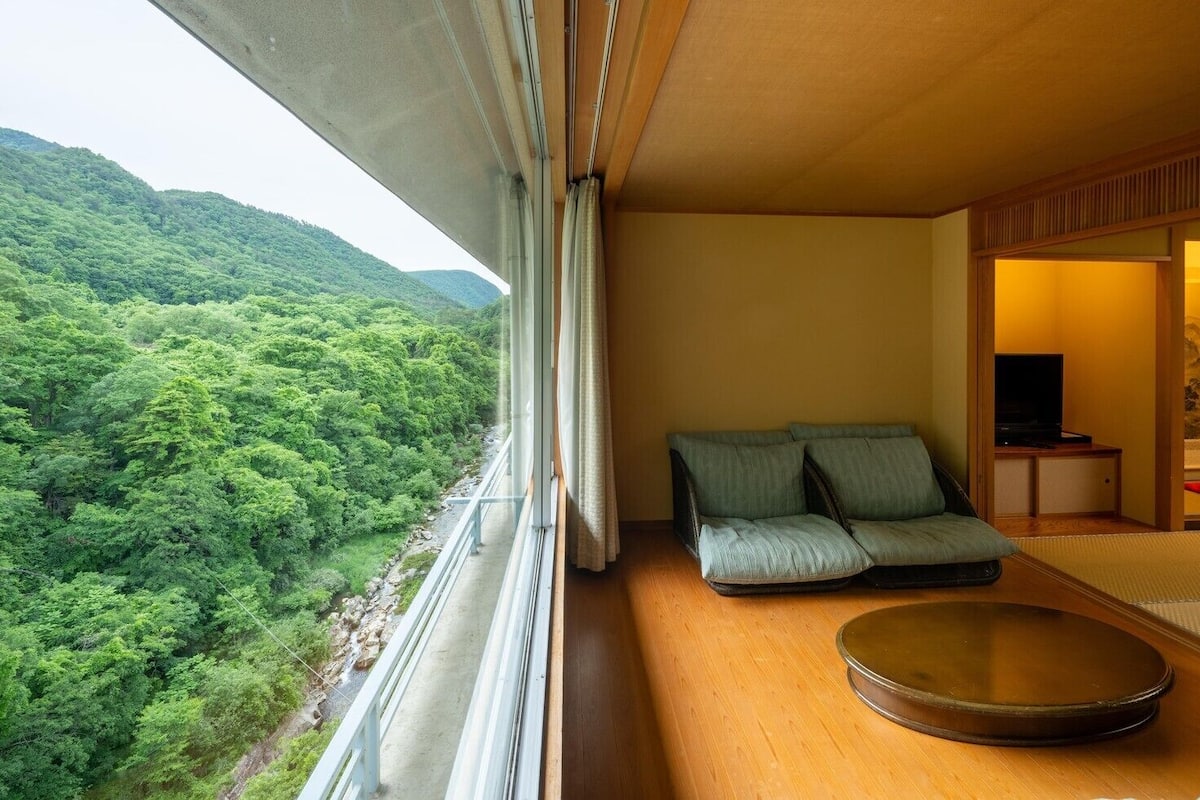 A room with a view of a forested hillside, a sofa, a tatami mat, and a small table with a television.
