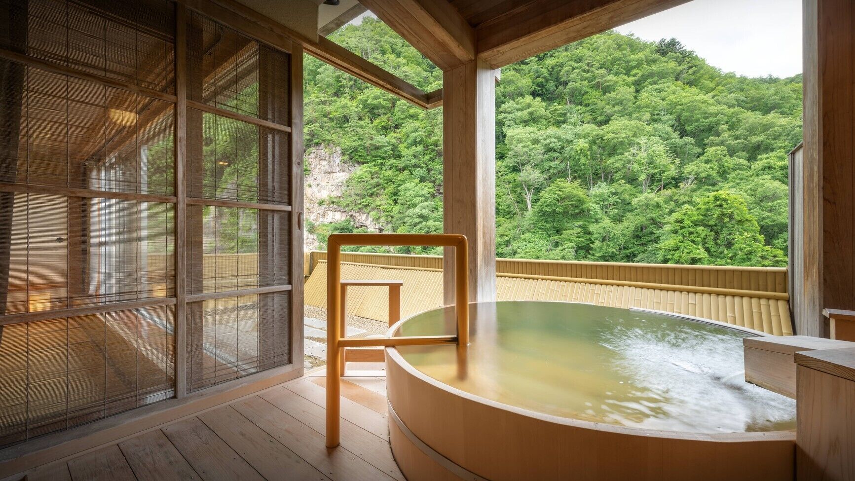 A hot spring bath area with wooden flooring, a wooden chair, and a view of a forested hillside.
