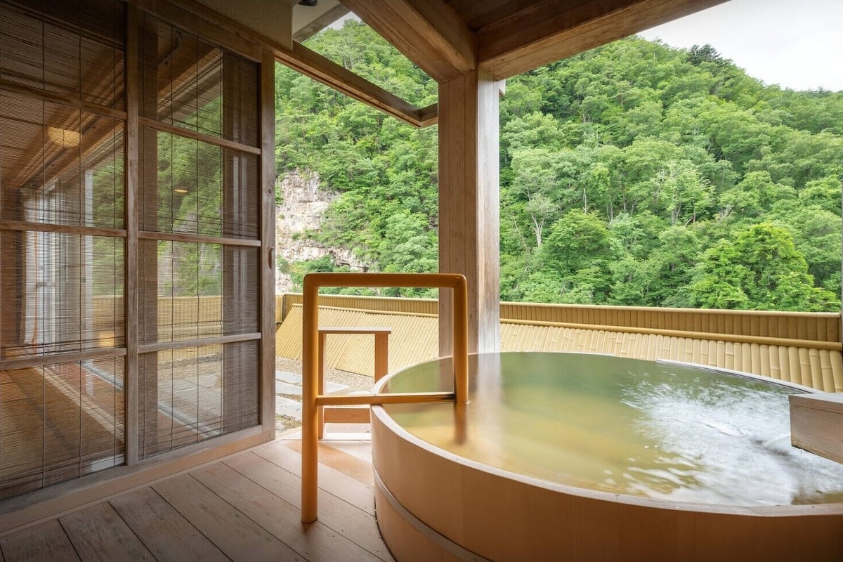 A hot spring bath area with wooden flooring, a wooden chair, and a view of a forested hillside.