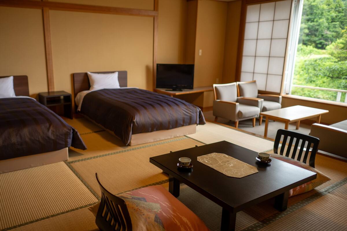 A hotel room with two beds, a TV, a table with a tea set, and a view of greenery outside.