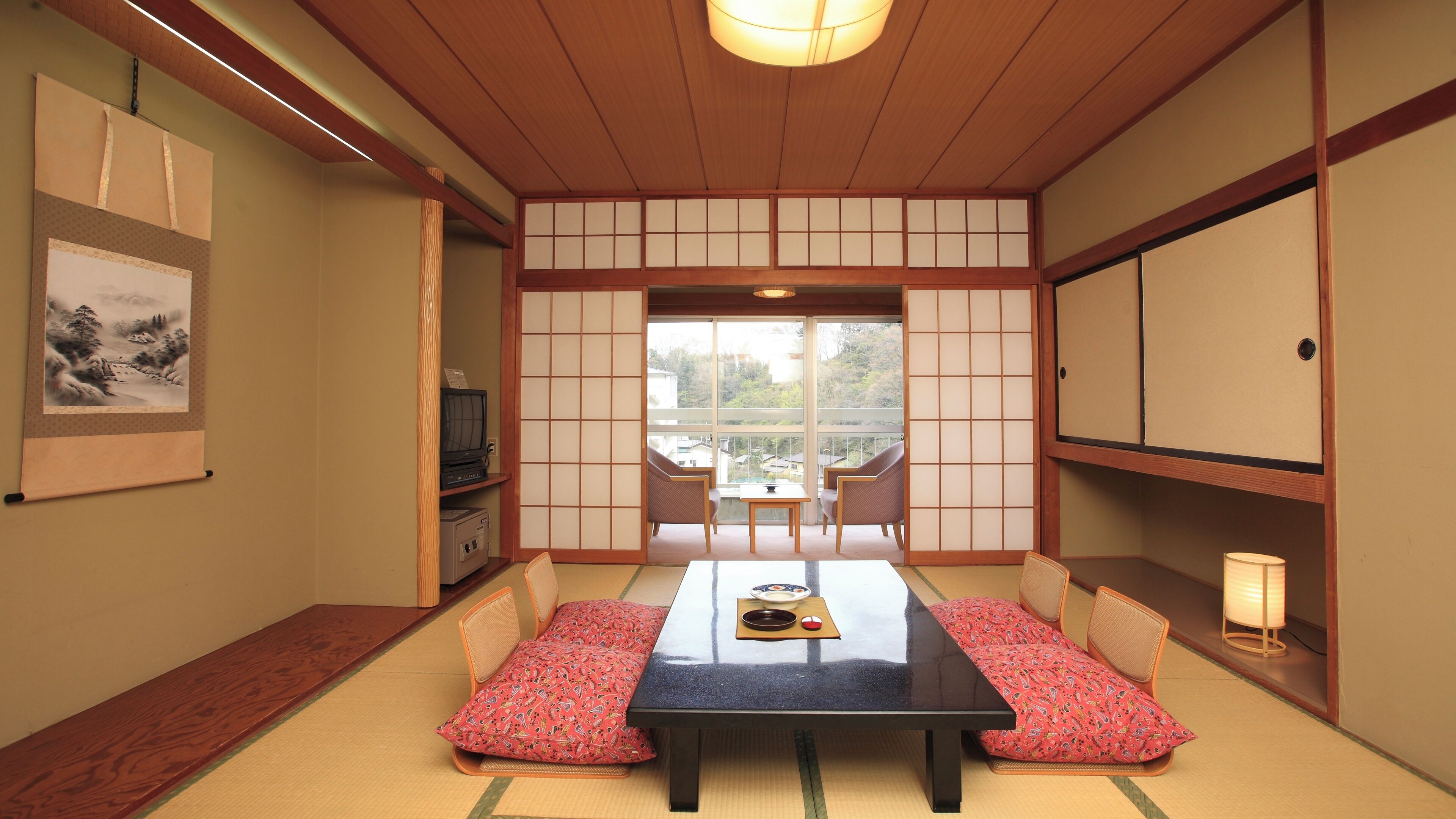 Ein traditioneller japanischer Raum mit Tatami-Boden, einem niedrigen Tisch, Kissen, Schiebetüren und einem Fernseher.