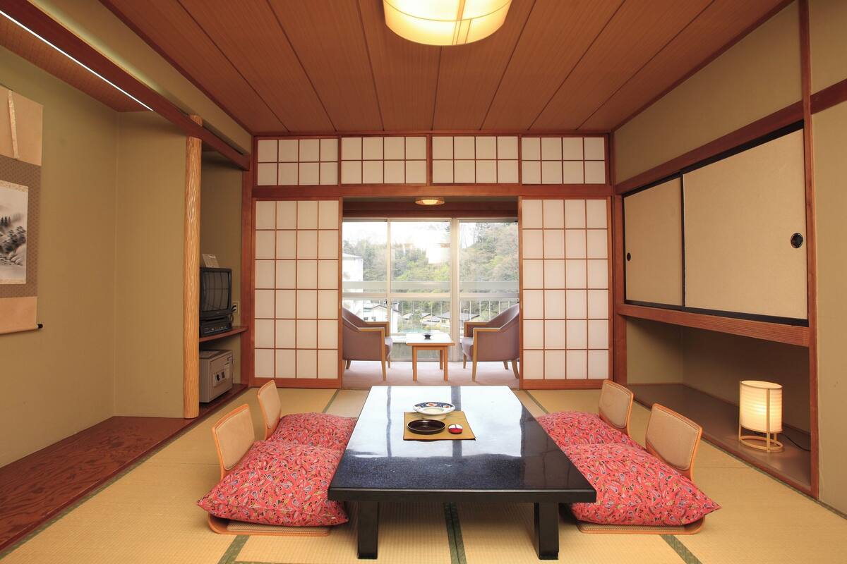 A traditional Japanese room with tatami flooring, a low table, cushions, sliding doors, and a television.