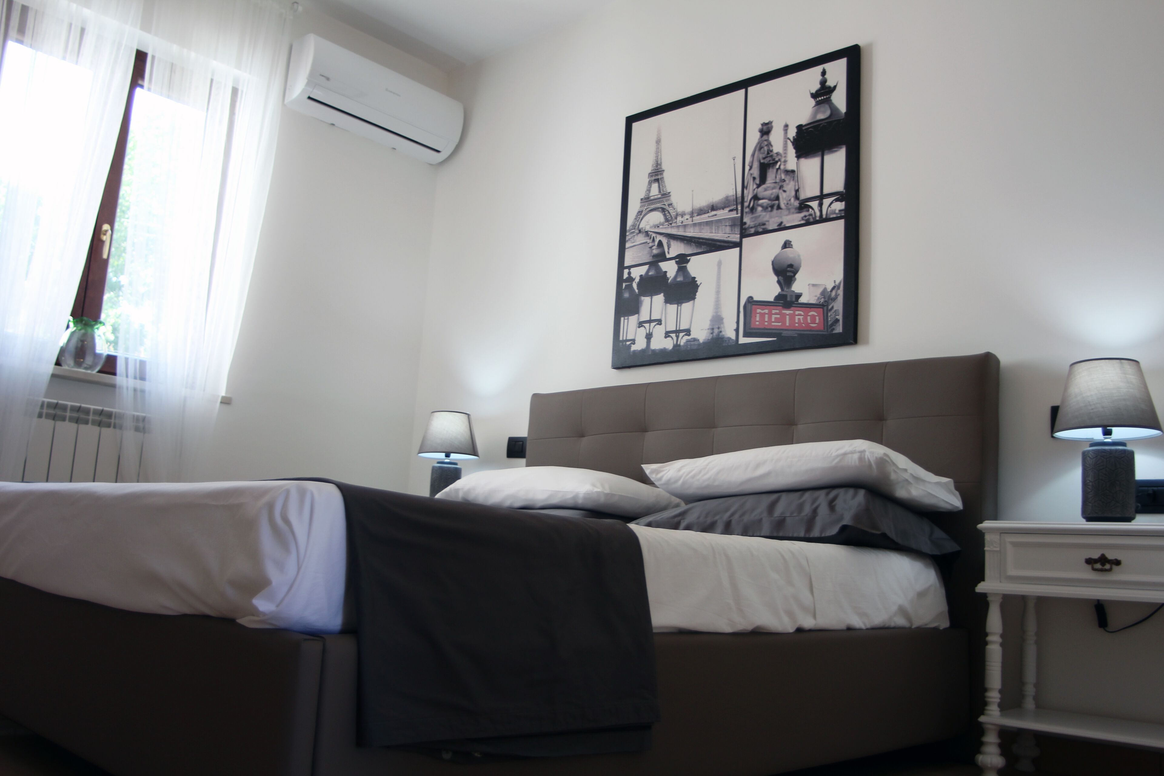 A hotel room with a bed, a nightstand with a lamp, a wall-mounted air conditioner, and a framed picture of Paris landmarks.