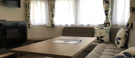 Uma sala de estar com sofá, mesa, TV e janelas com cortinas.