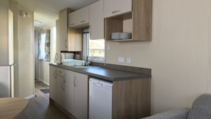 A compact kitchen with a dishwasher, a sink, and a countertop. There is a wooden cabinet above the sink and a small shelf with plates. - The Leas Beach Park (Norwich)
