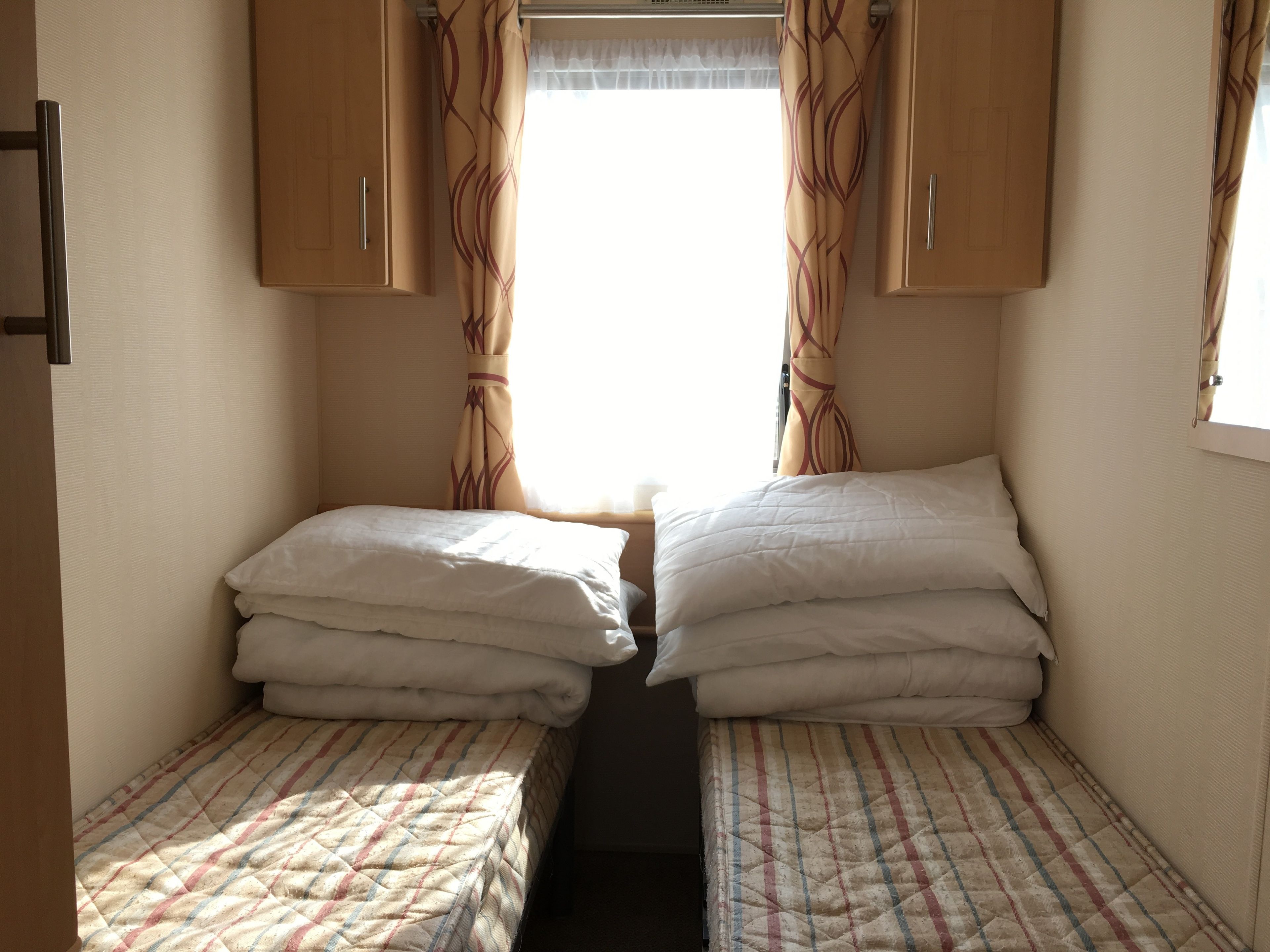 Une chambre compacte dans un camping-car, avec deux lits simples, une fenêtre avec des rideaux et des placards en bois.