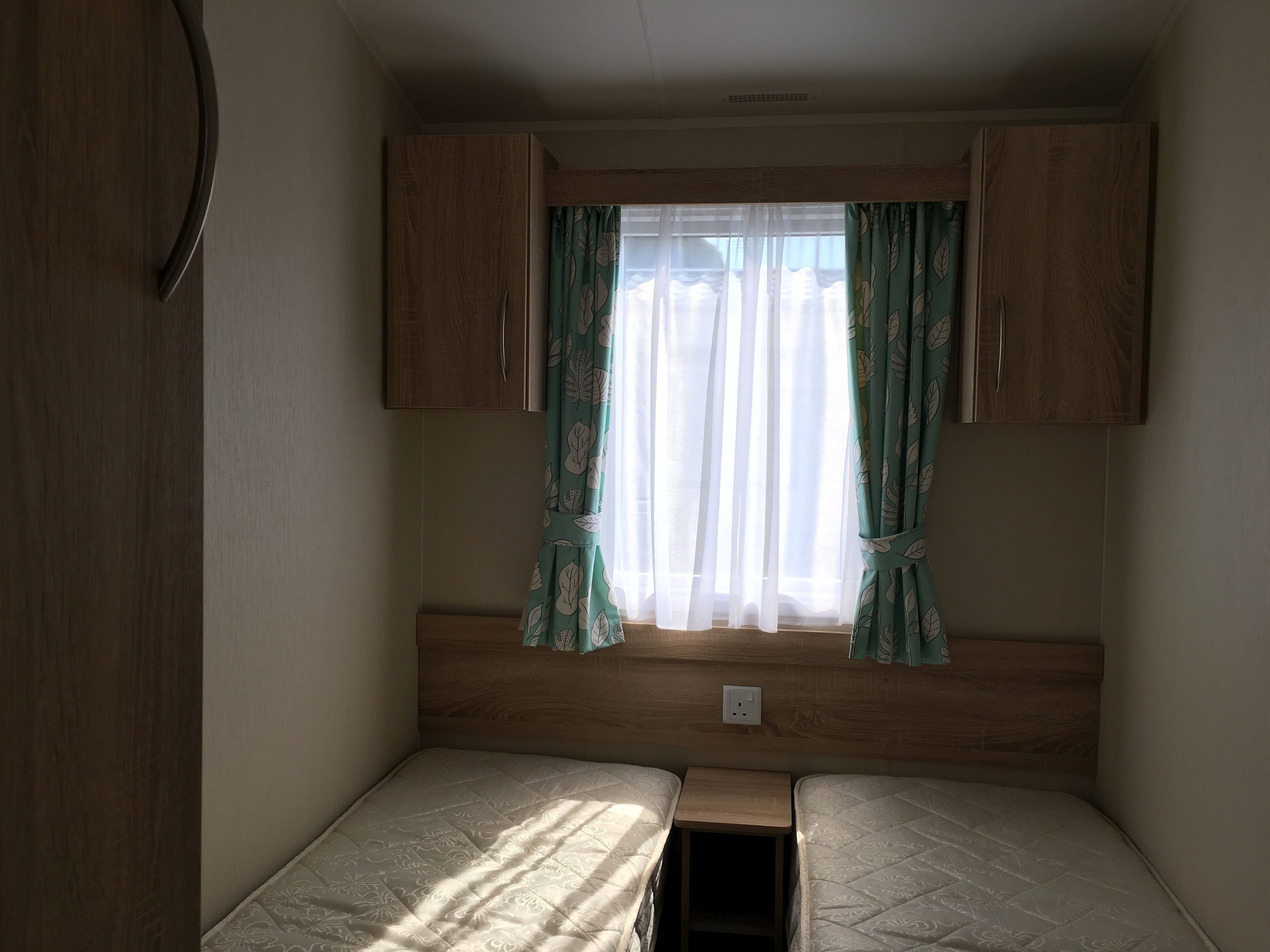 A small, enclosed room with two single beds, a window with sheer curtains, and wooden cabinets.