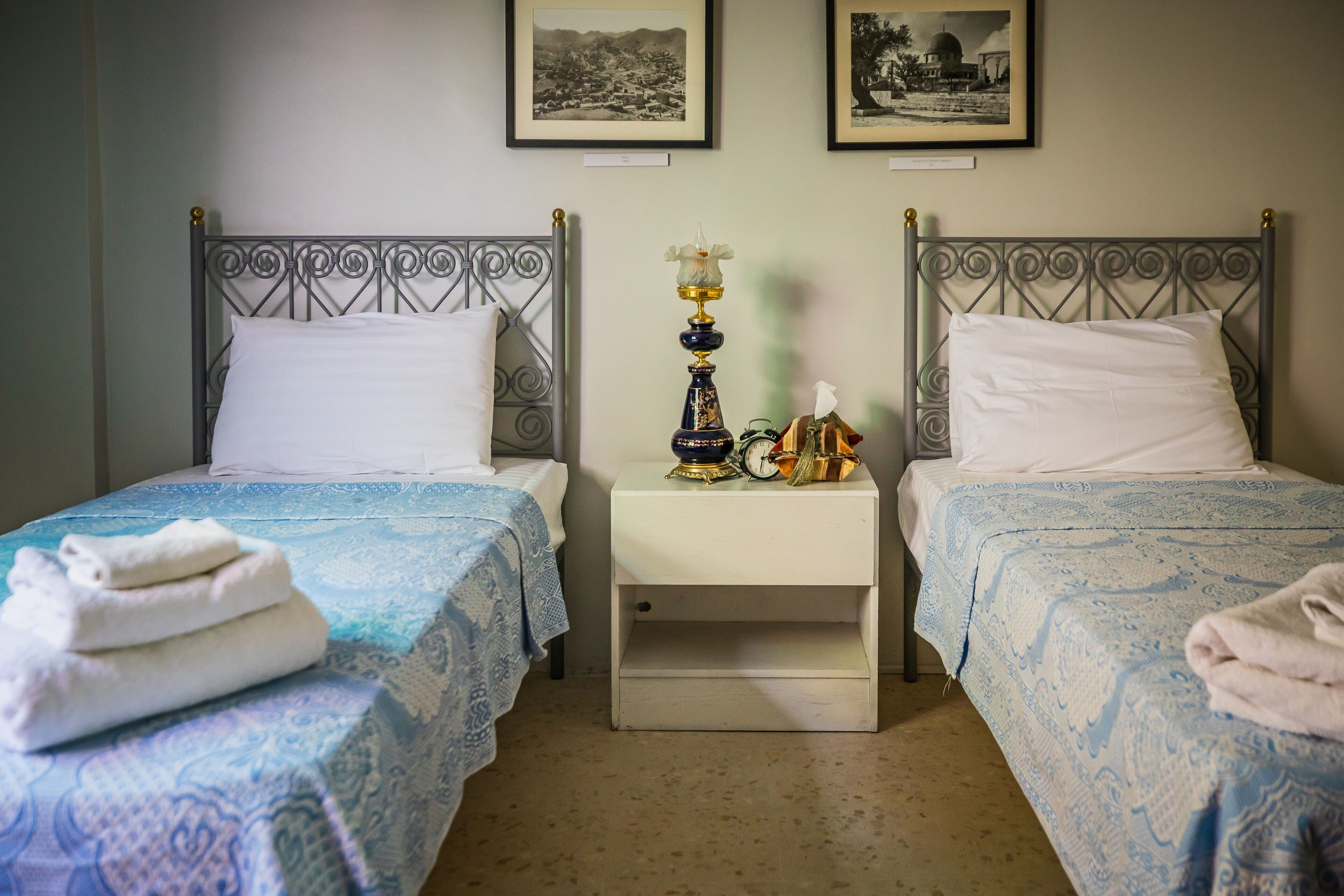 Un dormitorio con dos camas, cada una con una colcha estampada y almohadas blancas. Entre las camas hay una mesita de noche con una lámpara decorativa y un pequeño reloj.