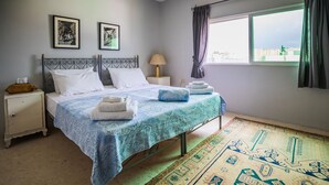 Una cama bien hecha con una colcha azul y blanca estampada, una lámpara de noche y vistas de la ciudad a través de una ventana.