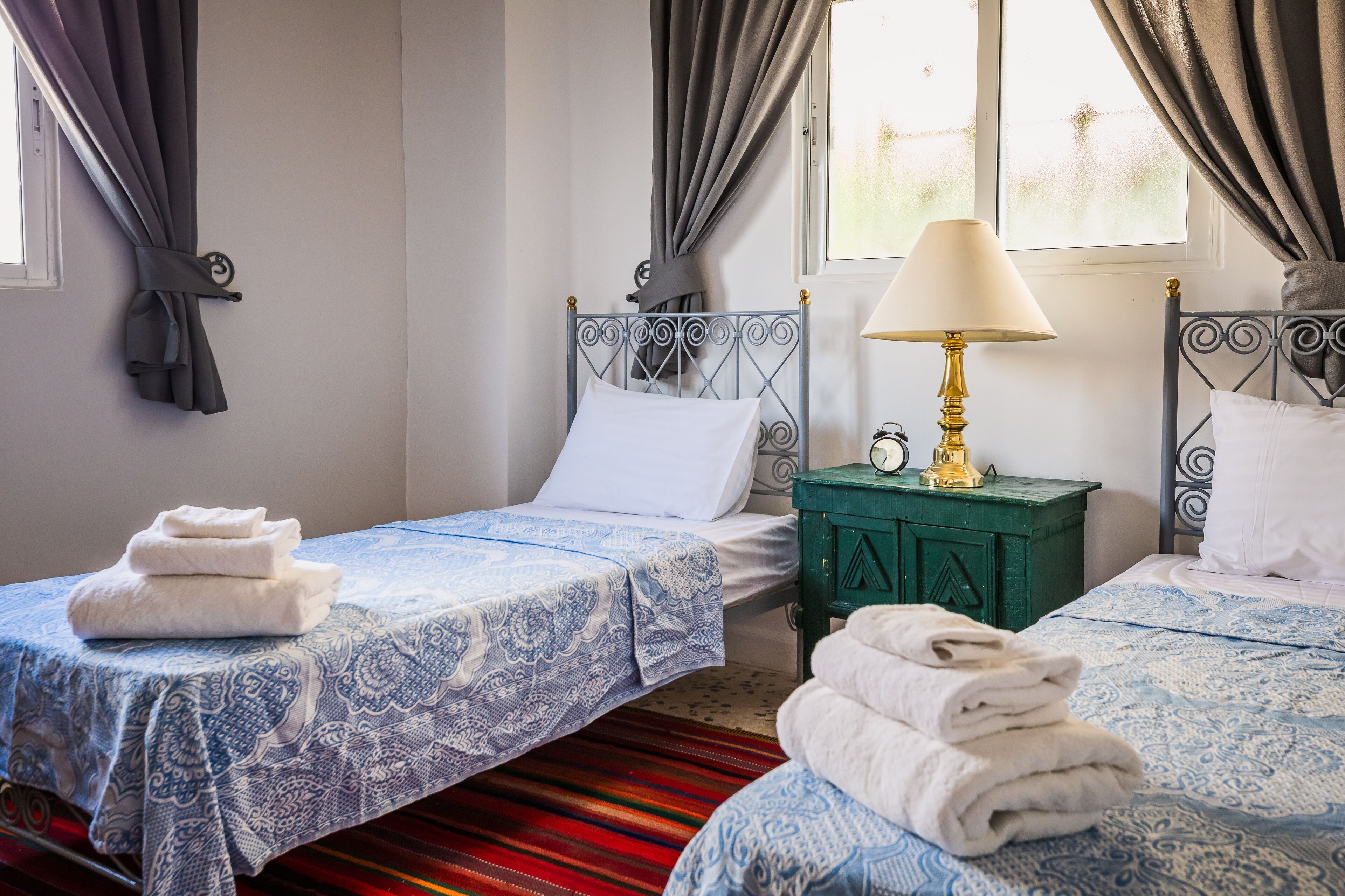 A hotel room with two beds, a green nightstand with a lamp, and a window with curtains.