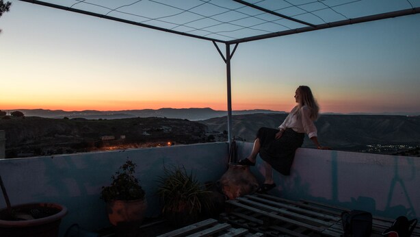 Una mujer sentada en un balcón con vistas a un paisaje urbano al atardecer.