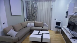 A modern living room with a sectional sofa, a coffee table, a TV, and a fan. - Doka Apartments (Sarandë)