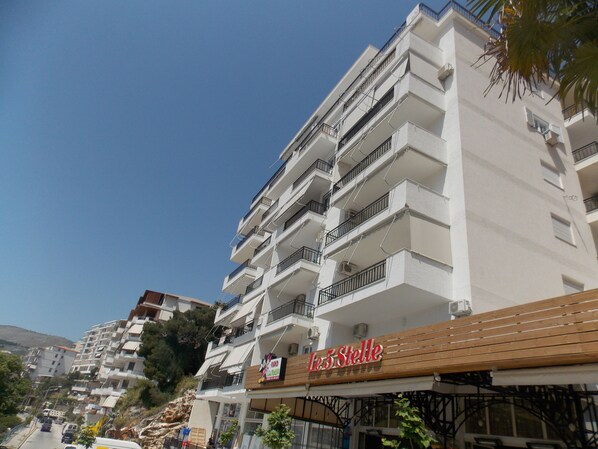 A multi-story building with balconies, a restaurant named "Le 5 Stelle" on the ground floor, and a clear blue sky in the background. - Doka Apartments (Sarandë)