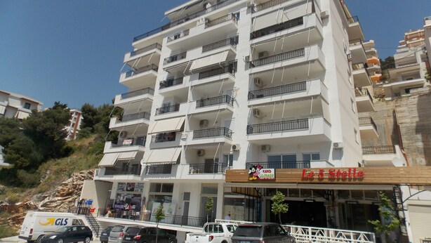 A multi-story building with balconies, a restaurant named "Le 5 Stelle" on the ground floor, and several parked vehicles including a van and cars. - Doka Apartments (Sarandë)