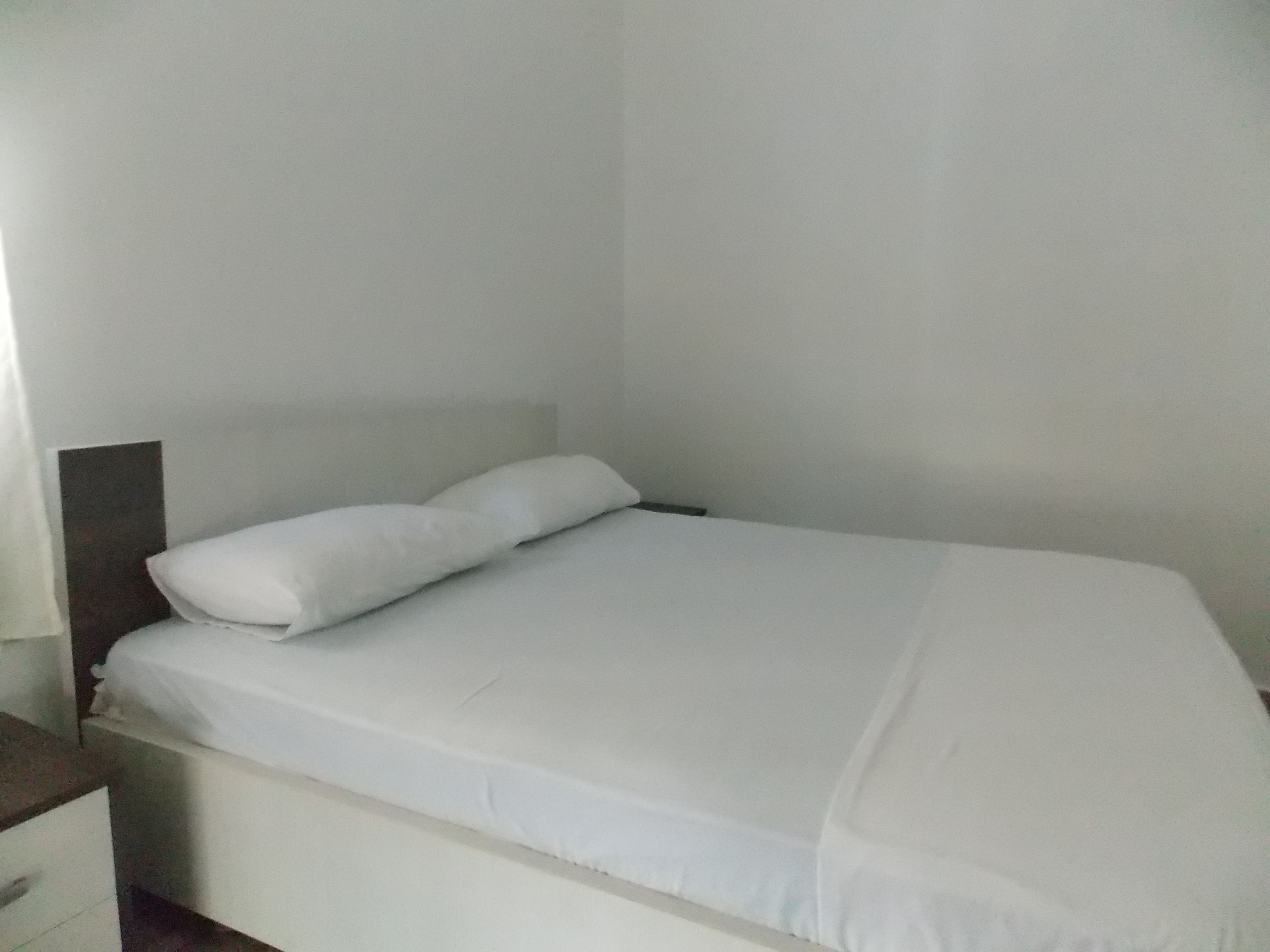 A single-bed room with a white bedspread and pillows, a wooden headboard, and a nightstand with a drawer.