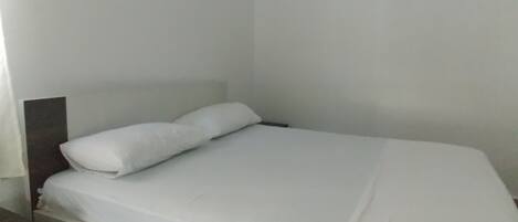 A single-bed room with a white bedspread and pillows, a wooden headboard, and a nightstand with a drawer.