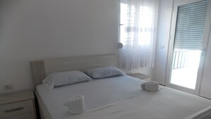 A neatly made bed with white linens and pillows, a wooden headboard, a bedside table with a lamp, and a window with sheer curtains. - Doka Apartments (Sarandë)