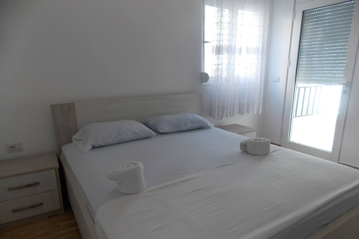 A neatly made bed with white linens and pillows, a wooden headboard, a bedside table with a lamp, and a window with sheer curtains.