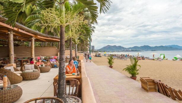 A beachside restaurant with outdoor seating, palm trees, and a view of the beach and mountains. - Hanka Star Hotel (Nha Trang)