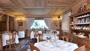 Un comedor rústico con mesas y sillas de madera, manteles blancos y una repisa con vajilla y una cesta de frutas.