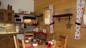 L’intérieur est en bois et comprend une table à manger pour quatre personnes, entourée de chaises en bois. Sur la table, on trouve un chemin de table rouge, un vase avec des fleurs rouges et un chandelier rouge. Un four à micro-ondes et une machine à café sont posés sur le comptoir.