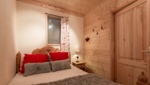 Une chambre à coucher aux murs lambrissés, avec un lit, des oreillers rouges et gris, une table de chevet avec une lampe et une fenêtre avec un rideau.