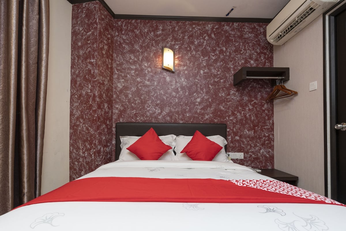 A hotel room with a bed, red and white bedding, a wall-mounted light, and an air conditioning unit.