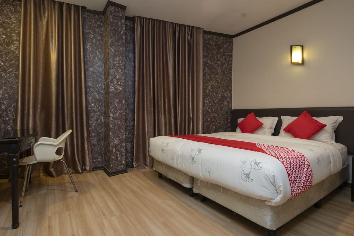 A hotel room with a double bed, two red pillows, a white bedspread, a red and white throw, a desk with a chair, and a wall-mounted light.