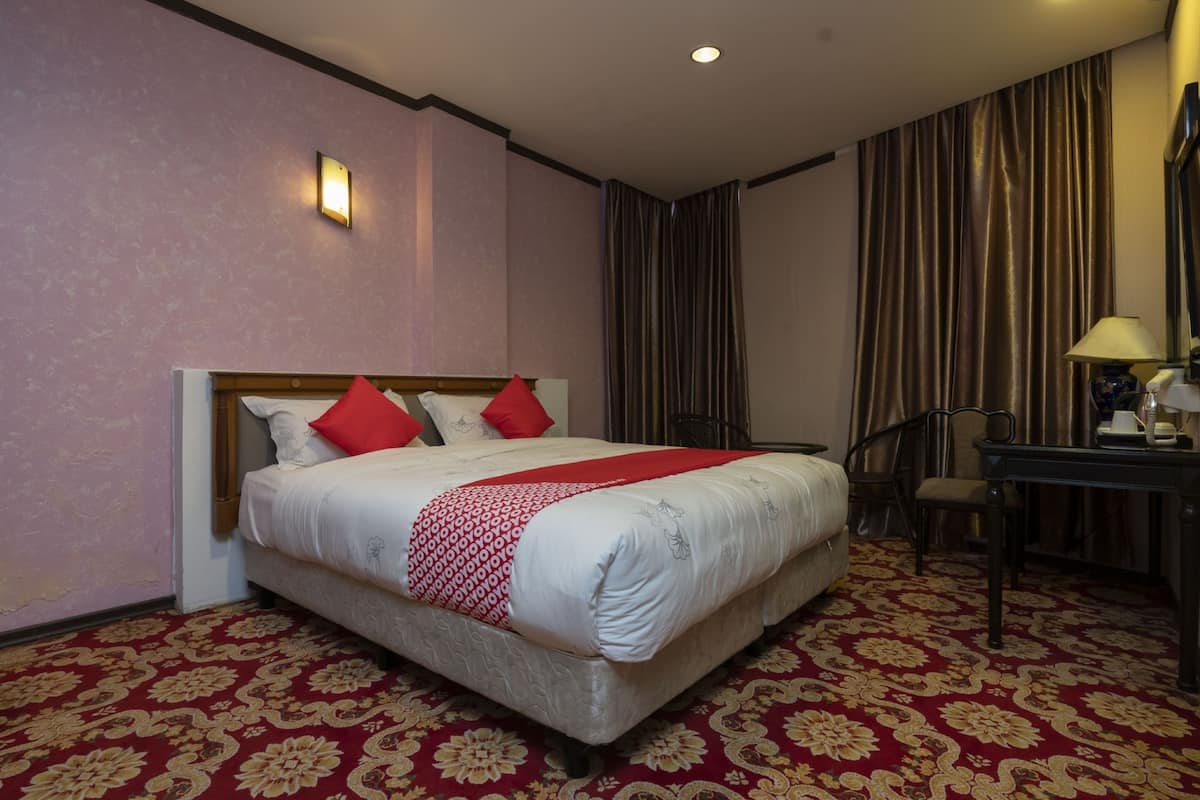 A hotel room with a large bed, red and white bedding, a red and gold carpet, a desk with a lamp, and a chair.