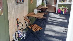 A covered outdoor seating area with wooden tables and chairs, a small table with a potted plant, and a wall-mounted light fixture.
