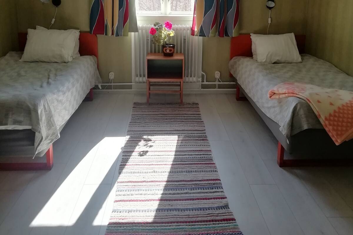 Une chambre avec deux lits, une fenêtre avec des rideaux, un radiateur et une petite table sur laquelle se trouve un vase.