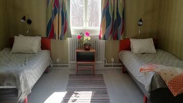 A bedroom with two beds, a window with curtains, a radiator, and a small table with a vase.
