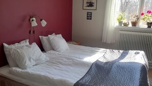 A bedroom with a bed, a nightstand, a wall-mounted lamp, a window with curtains, and a radiator.