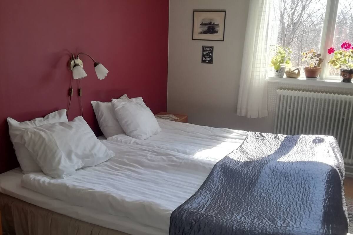 Une chambre à coucher comprenant un lit, une table de chevet, une lampe fixée au mur, une fenêtre avec des rideaux et un radiateur.