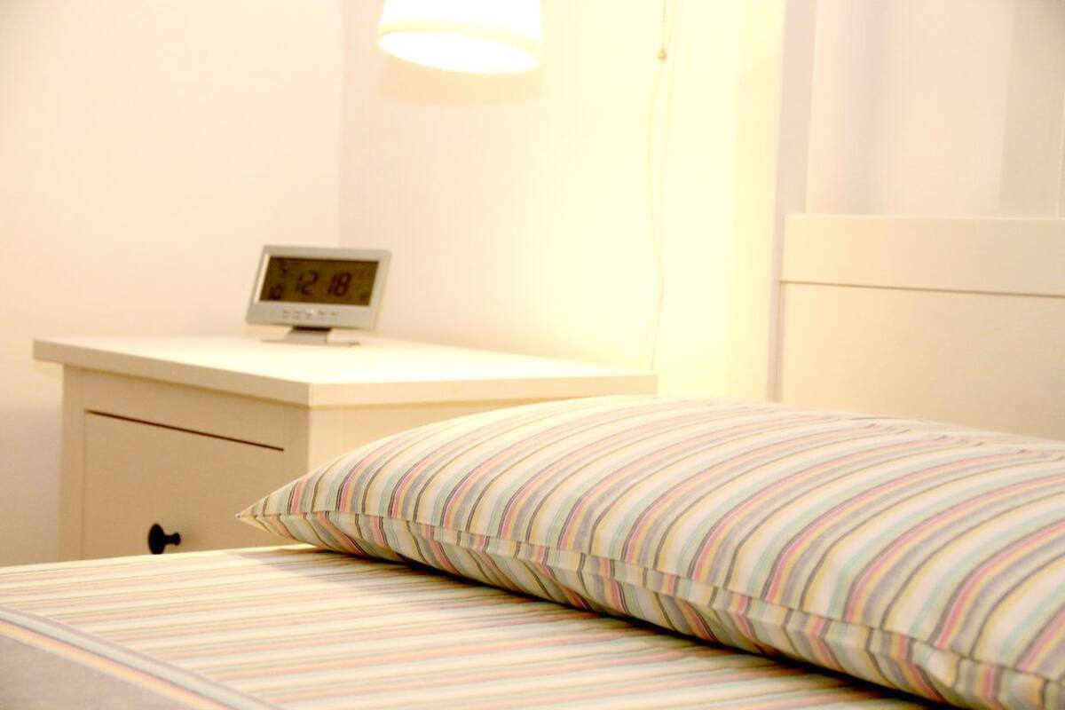 Una cama bien hecha, con un almohadón a rayas y una mesita de noche con un reloj digital.