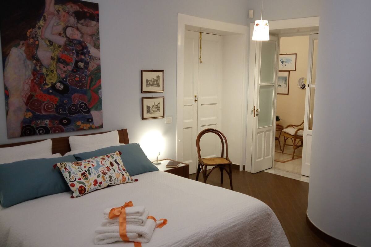 A hotel room with a bed, a chair, a desk, and a painting on the wall.