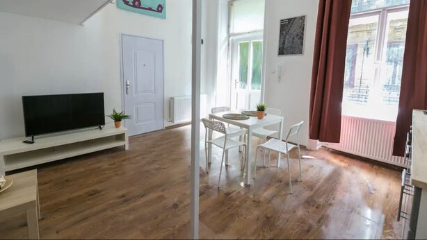 Ein Zimmer mit Fernseher auf einem Schrank, einem Esstisch mit Stühlen, einer weißen Tür und einem Fenster mit Vorhängen.